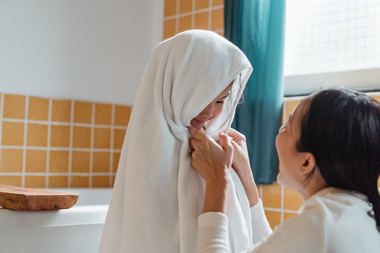 Cheerful ethnic mum wiping cute little daughter with big fluffy towel in modern bathroom at home